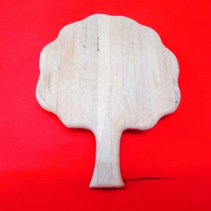 May include: A light-colored wooden cutting board in the shape of a tree. The tree-shaped board has a wide, irregular top and a narrow, rectangular handle. The board is made of vertical wooden planks and sits against a bright red background.