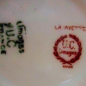 May include: Close-up of a white porcelain surface with two distinct markings. One marking is in green and reads "U.C. Limoges France". The other is a red emblem with the words "LA FAYETTE" and "U.C. Limoges FRANCE".