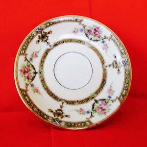 May include: A vintage porcelain saucer with a white center and a decorative border. The border features floral bouquets in pink and purple, with accents of green, gold, and blue. The saucer has a gold rim and intricate detailing.