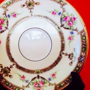 May include: A vintage porcelain saucer with a cream-colored base and a central white circle. The saucer is decorated with pink roses, blue accents, and a brown and gold geometric border. The rim is trimmed with gold.