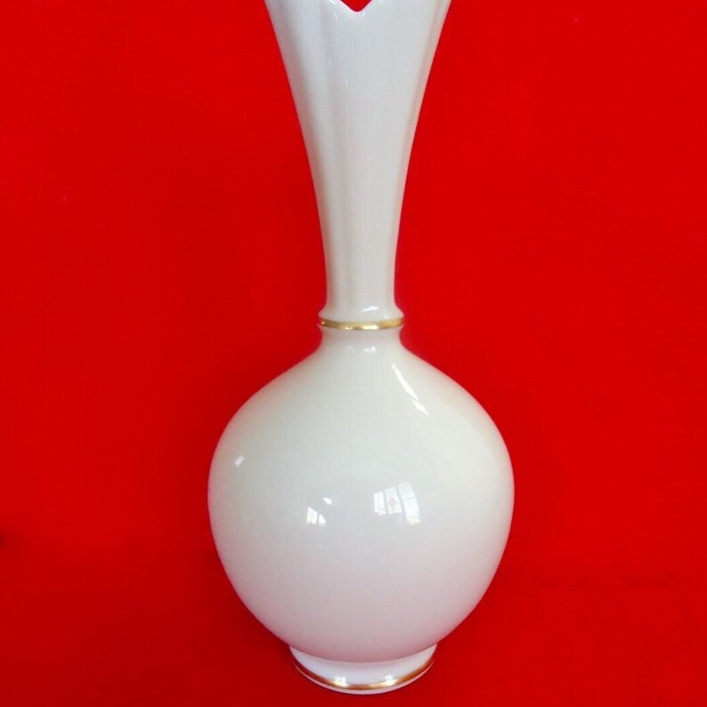 May include: A white ceramic vase with a bulbous base and a tall, fluted neck. The vase has a gold trim around the base and neck. The background is a solid red.