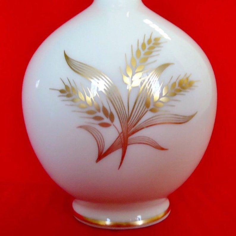 May include: A white, round vase with a gold wheat stalk design. The vase has a small, rounded base with a gold trim. The wheat design is detailed with gold and brown accents, set against the smooth, white surface of the vase.