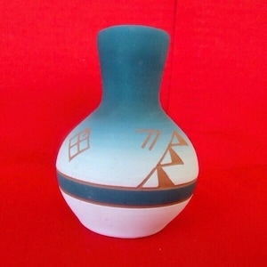 May include: A small, turquoise and white vase with a narrow neck and rounded body. The vase features brown geometric designs and a brown band near the base. The background is a solid red.