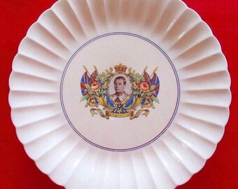 Op de afbeelding: Witte keramische plaat met een geschulpte rand. Het midden toont een portret in een wapen, geflankeerd door vlaggen en bloemmotieven. Een blauwe rand omcirkelt het wapen. De plaat staat op een rode achtergrond.