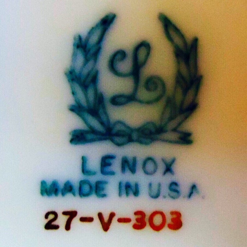 May include: Close-up of a white ceramic surface with a blue and green emblem featuring the letter "L" inside a wreath. Below the emblem, the text "LENOX MADE IN U.S.A." is printed in blue, with "27-V-303" in red.