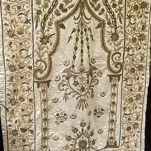 Peut inclure: Un textile rectangulaire blanc cassé orné de broderies dorées complexes. Le motif présente une arche centrale, un élément en forme de lanterne suspendue et des motifs floraux denses avec des croissants de lune et des étoiles.