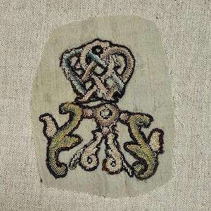 May include: An antique embroidered patch with a complex design. The patch features a central knot-like motif in shades of blue, tan, and brown, with stylized green and yellow elements below. The patch is on a light green fabric.