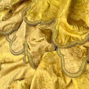 May include: Close-up of a gold fabric with a floral pattern and a gold trim. The fabric is folded and draped, showing the texture and detail of the trim.
