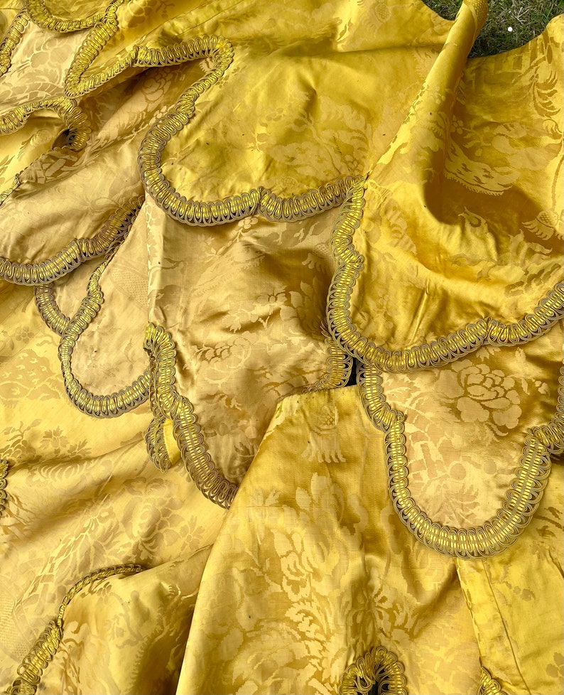 May include: Close-up of a gold fabric with a floral pattern and a gold trim. The fabric is folded and draped, showing the texture and detail of the trim.