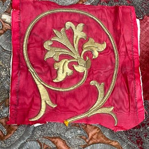 May include: A square piece of red fabric featuring a central gold metallic thread embroidery in a floral motif enclosed within a circular gold border. The red fabric is layered over a larger brown textile with intricate silver metallic embroidery.