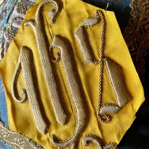 May include: A close-up of a yellow fabric with gold embroidery. The embroidery forms ornate, stylized letters. The fabric is bordered by gold trim and rests on a patterned blue fabric.