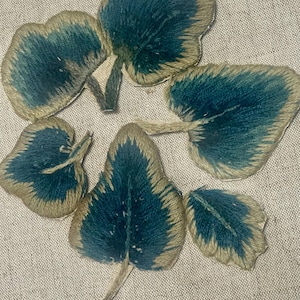 May include: Five embroidered leaf-shaped patches featuring a deep teal center and a light beige border. Each patch has a distinct stem and a textured, stitched appearance with visible thread details throughout the leaf surface.