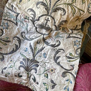以下が含まれることがあります： クリーム色の生地に、精巧な花の刺繍が施されています。刺繍はグレー、ブルー、グリーン、ピンクのアクセントが特徴です。このパターンには、様式化された花、葉、スクロール要素が含まれており、ヴィンテージな美しさを生み出しています。