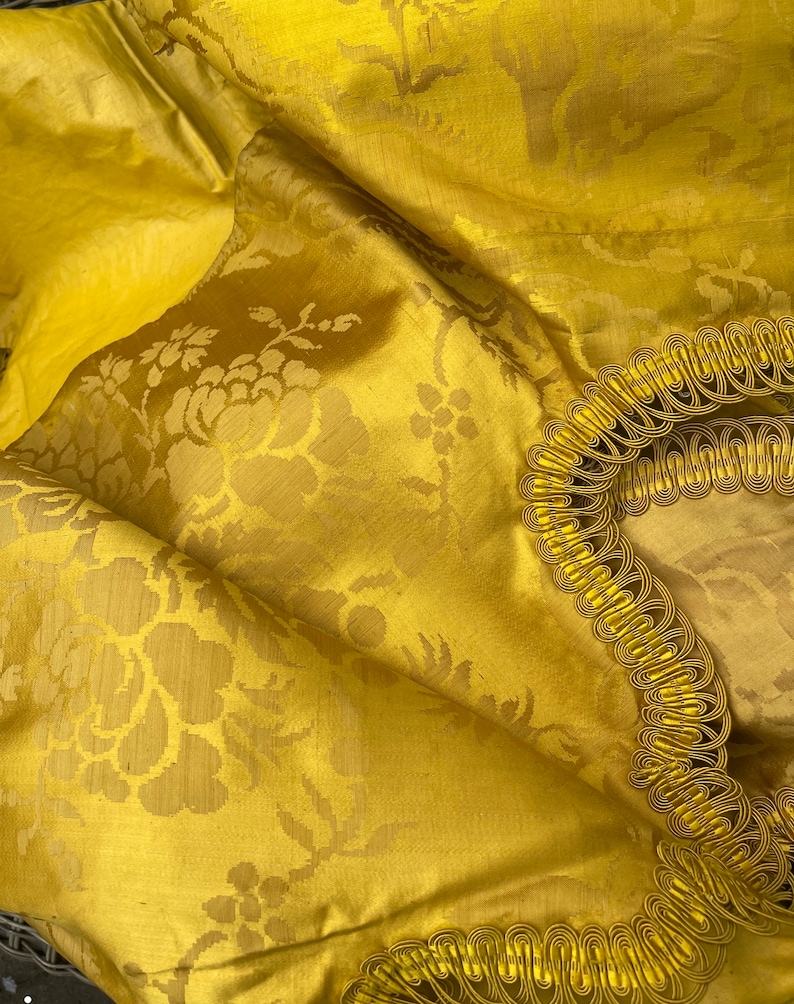 May include: A close-up of a yellow fabric with a floral pattern. The fabric has a gold trim around the edges.