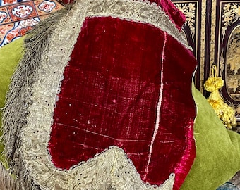 May include: A deep red velvet textile with a gold lace trim and fringe. The textile is draped over a green cushion. The gold trim has a delicate, openwork design. The fringe is a muted gold color.
