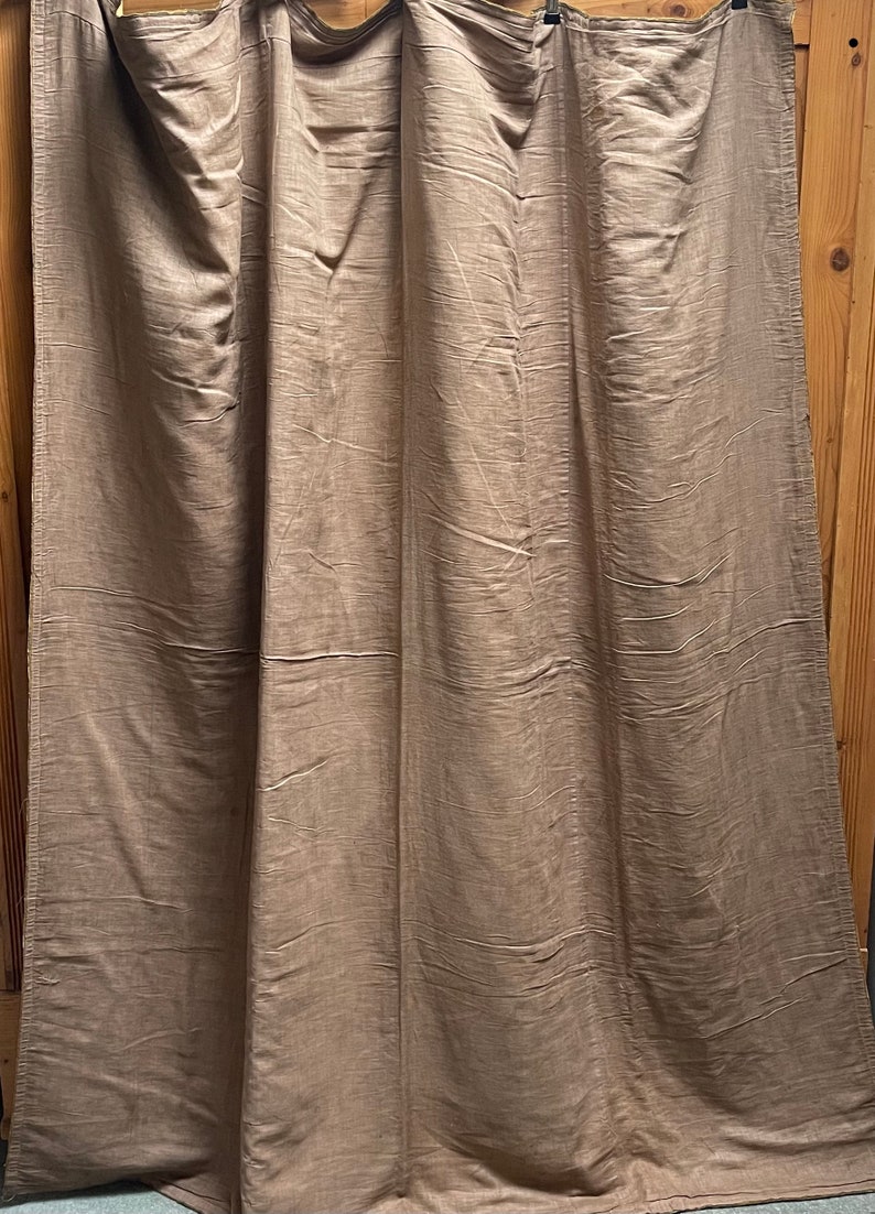 May include: A set of three brown linen curtains hanging on a wooden rod. The curtains are made of a soft, natural fabric and have a subtle texture.