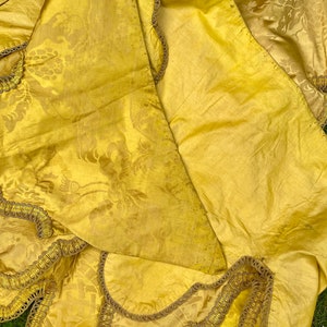 May include: A yellow fabric with a floral pattern and gold trim. The fabric is folded in half and the trim is visible on both sides.