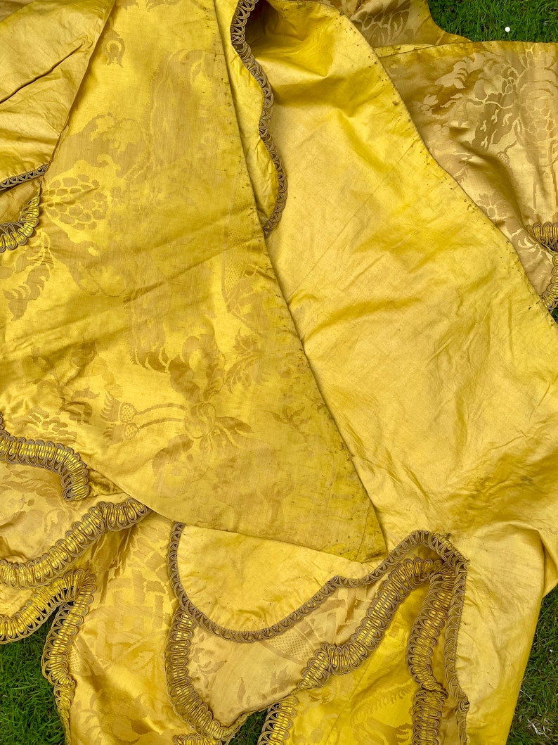 May include: A yellow fabric with a floral pattern and gold trim. The fabric is folded in half and the trim is visible on both sides.