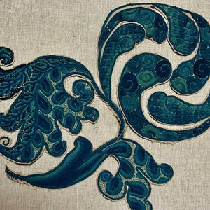 May include: Close-up of an embroidered design featuring teal and dark blue floral patterns on a beige linen background. The design includes stylized leaves and swirling shapes, showcasing detailed stitching.