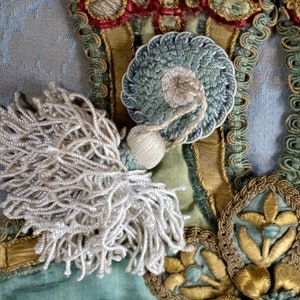 May include: A white tassel with a blue and white circular embellishment. The tassel is attached to a fabric background with gold and green embroidery.