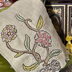 Może przedstawiać: Fragment antycznej tkaniny z haftowanym wzorem kwiatowym. Haft przedstawia różowe, żółte i zielone kwiaty i liście na beżowym tle. Projekt zawiera stylizowany węzeł u podstawy kompozycji kwiatowej.