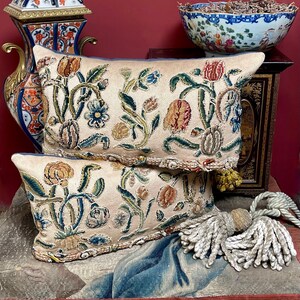 May include: Two decorative rectangular pillows with floral embroidery in beige, blue, green, and orange. The pillows have a light blue border and are stacked. A tassel with a cream-colored rope is visible.