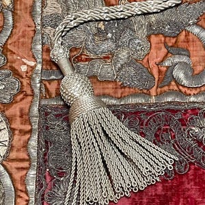 May include: A silver metallic tassel with a braided cord and a rounded knot head, resting on a textile surface featuring intricate silver thread embroidery on a background of deep orange and dark red velvet fabric with ornate floral patterns.