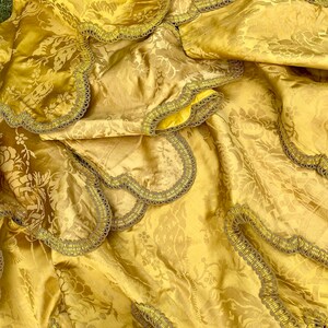 May include: A gold fabric with a floral pattern and a decorative trim. The fabric is folded and draped, showing the texture and detail of the trim.