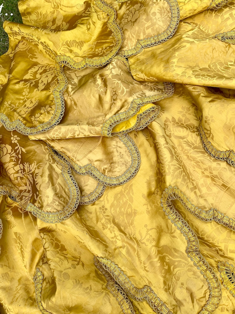 May include: A gold fabric with a floral pattern and a decorative trim. The fabric is folded and draped, showing the texture and detail of the trim.