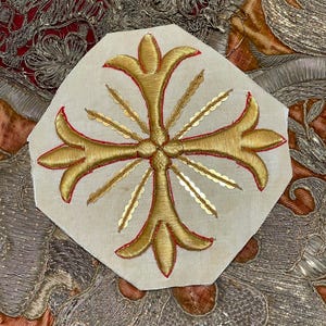 May include: A gold embroidered cross with a red outline on a white fabric background. The cross has a stylized design with a sunburst of rays emanating from the center.