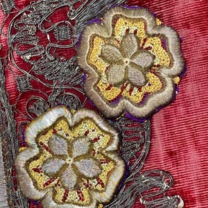 May include: Two embroidered floral motifs featuring metallic silver and gold thread on a deep red velvet fabric. The flowers have five petals with intricate stitching, layered over silver wire filigree and purple fabric accents.