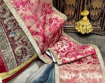 Puede incluir: Primer plano de un textil rosa y dorado con un estampado floral. El textil tiene un ribete y flecos dorados. El textil está doblado sobre un tapiz con un diseño azul y blanco. Una borla dorada está en el fondo.