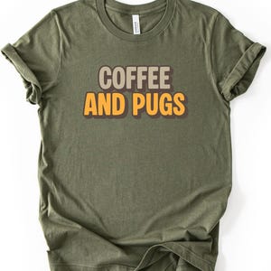 May include: Military green t-shirt with rolled sleeves. The shirt features the text "COFFEE AND PUGS" in a layered design with brown and orange lettering. The shirt is made of a soft material.