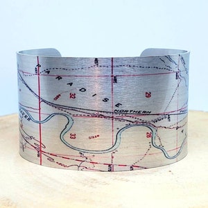 May include: A silver metal cuff bracelet with a vintage map design. The map shows a river, roads, and numbered sections. The bracelet is a great way to show your love for maps and travel.