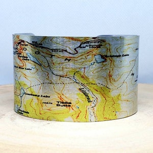Pode incluir: Uma pulseira de punho de metal prateado com um design de mapa topográfico em amarelo, verde e castanho. O mapa inclui curvas de nível, nomes de lugares e indicações de elevação. A pulseira é exibida numa superfície de madeira.