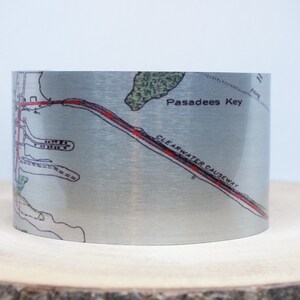 May include: A silver metal cuff bracelet with a map of Clearwater Causeway and Pasadees Key printed on it. The map is in black and white with red lines indicating the road.