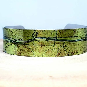 May include: A green and brown topographical map printed on a metal cuff bracelet. The map shows a river and the text "Kennebec River" and "Northern Railroad".