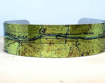May include: A green and brown topographical map printed on a metal cuff bracelet. The map shows a river and the text "Kennebec River" and "Northern Railroad".