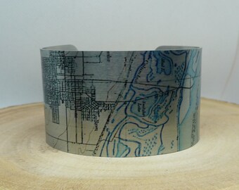 May include: A silver metal cuff bracelet with a vintage map design. The map features a coastal area with blue water and land features.