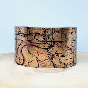 May include: A metal cuff bracelet with a vintage map design featuring the text "Branson" and "Big Pister" printed on it.