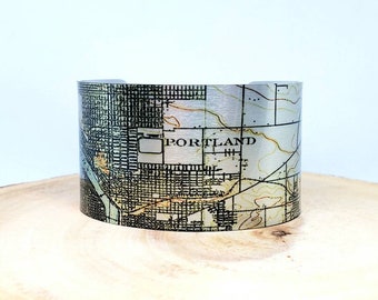 May include: A silver metal cuff bracelet with a vintage map of Portland, Oregon printed on it. The map is in shades of green, brown, and yellow.