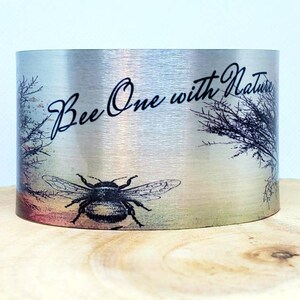 May include: A silver metal cuff bracelet with a black and white image of a bee and the text "Bee One with Nature".