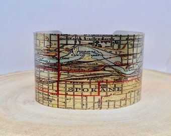 May include: A silver metal cuff bracelet with a vintage map of Spokane, Washington printed on it. The map is in shades of brown, tan, and red. The text "SPOKANE" is printed in red on the map.