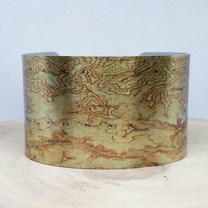 May include: A gold-toned metal cuff bracelet with a vintage map design. The map features brown and green lines and markings.