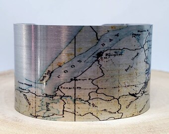 May include: A silver metal cuff bracelet with a vintage map of a coastal area printed on it. The map shows the names of various locations, including "North Bay", "Coos Head", and "North Spit".