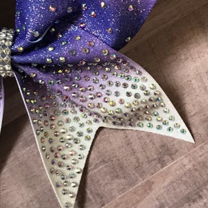 May include: A purple and white ombre cheer bow with iridescent rhinestones. The bow is made of glitter fabric and has a pointed shape.