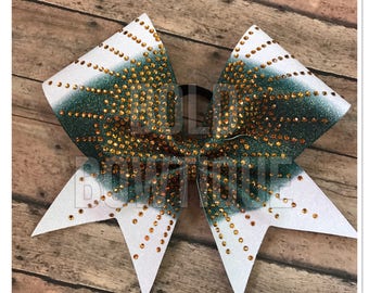 May include: A large, glittery cheerleading bow with a white and green ombre design. The bow is embellished with numerous gold-colored rhinestones. The bow is on a black elastic band, perfect for cheerleading or dance.