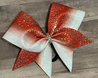 May include: A large orange and white ombre cheer bow with rhinestones. The bow is tied with a black elastic band.