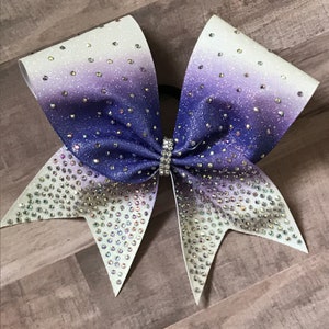 May include: A large purple and white cheer bow with iridescent glitter and rhinestones.