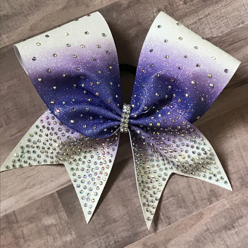 May include: A large purple and white cheer bow with iridescent glitter and rhinestones.